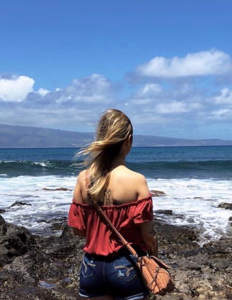 A woman in a red off-the-shoulder top and denim shorts stands on rocky terrain by the ocean, gazing at the waves under a bright blue sky with scattered clouds.
